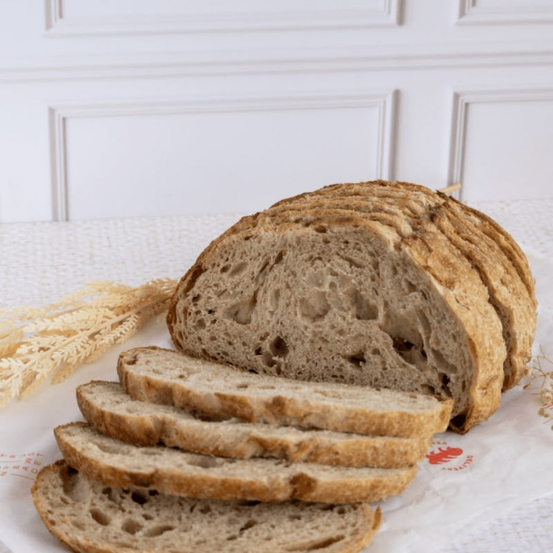 Hogaza de pan de masa madre en rebanadas sobre papel de panadería, con espigas decorativas al lado, disponible en Nutribody en variedades con semillas, arándano y nuez, dátil y romero, o aceituna negra.